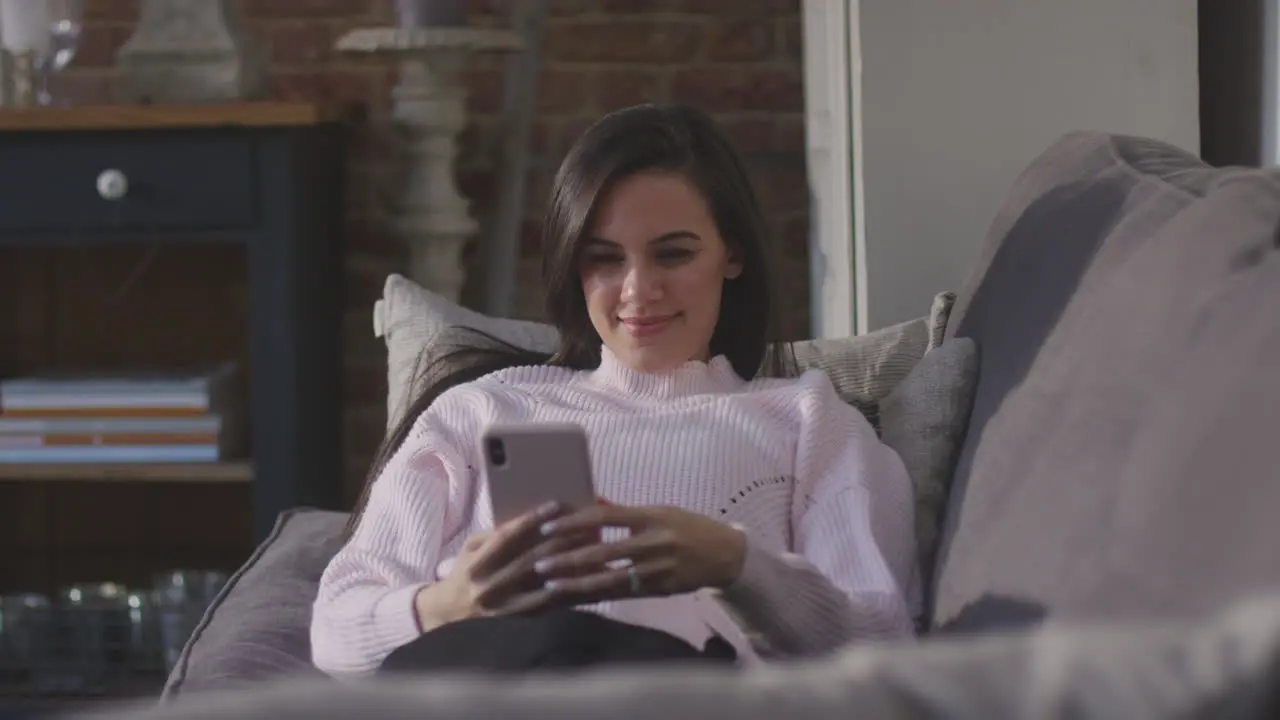 Smiling Woman Relaxing On Sofa At Home Checking Social Media On Mobile Phone