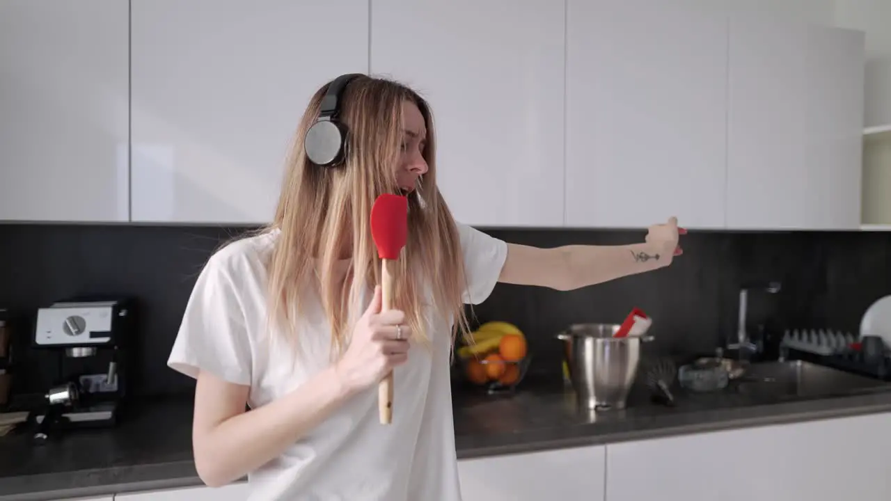 Young Woman In Headphones Dancing And Pretending Singing In Kitchen At Home