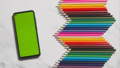 Coloured Pencils Arranged In A Symmetrical Pattern With Green Screen Mobile Phone On White Marble Background 