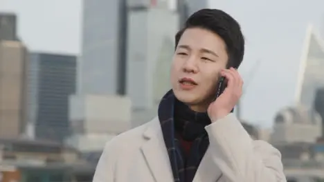 Young Asian Businessman Answering Call On Mobile Phone With London City Skyline In Background