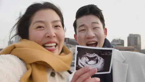 Pregnant Young Asian Couple In London UK Making Video Call Home On Phone Showing Sonogram Of New Baby To Family 1