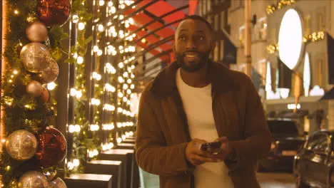 Man Walking Along Street On Christmas Shopping Trip To London Looking At Mobile Phone