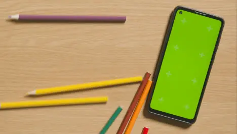 Multi-Coloured Pencils Rolling Down Wooden Background Next To Green Screen Mobile Phone