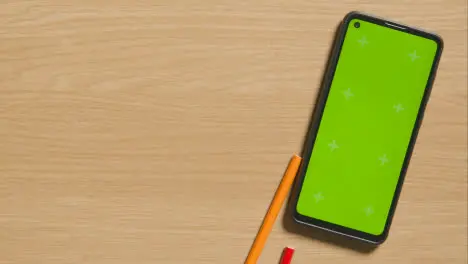 Multi-Coloured Pencils Rolling Down Wooden Background Next To Green Screen Mobile Phone 1