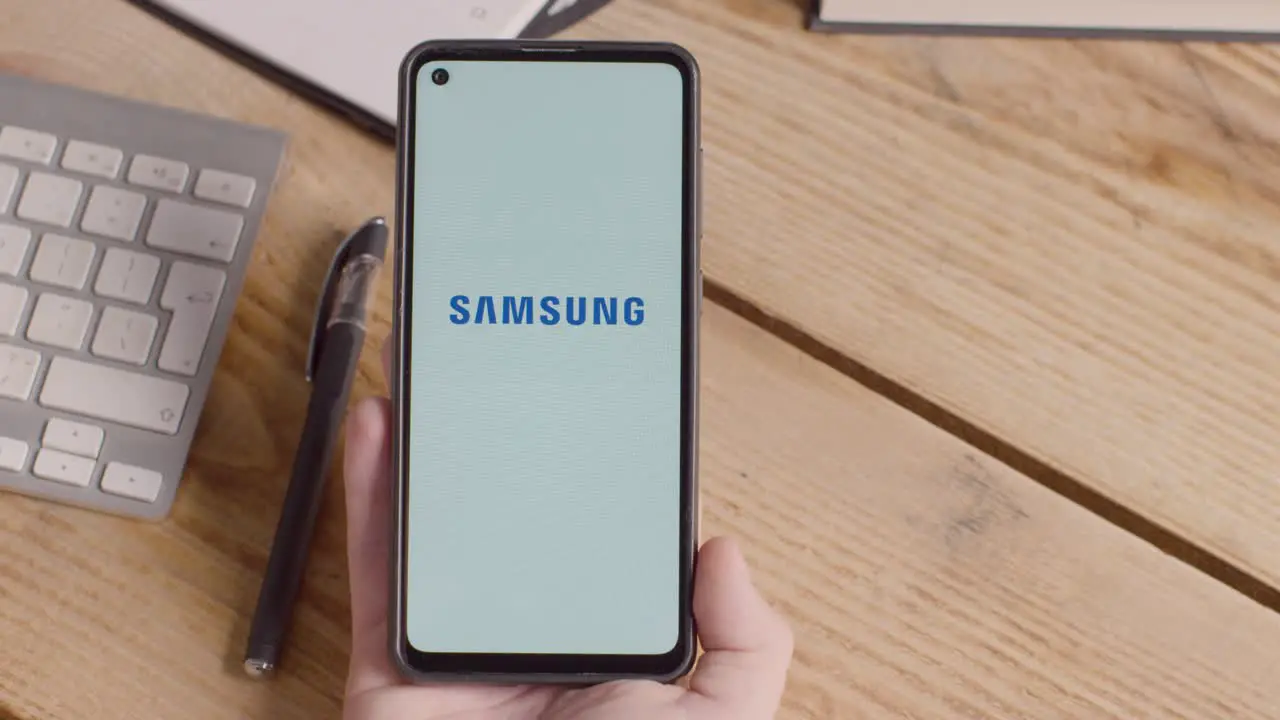 Person Holding Mobile Phone On Desk With Screen Showing Brand Logos For Apple And Samsung