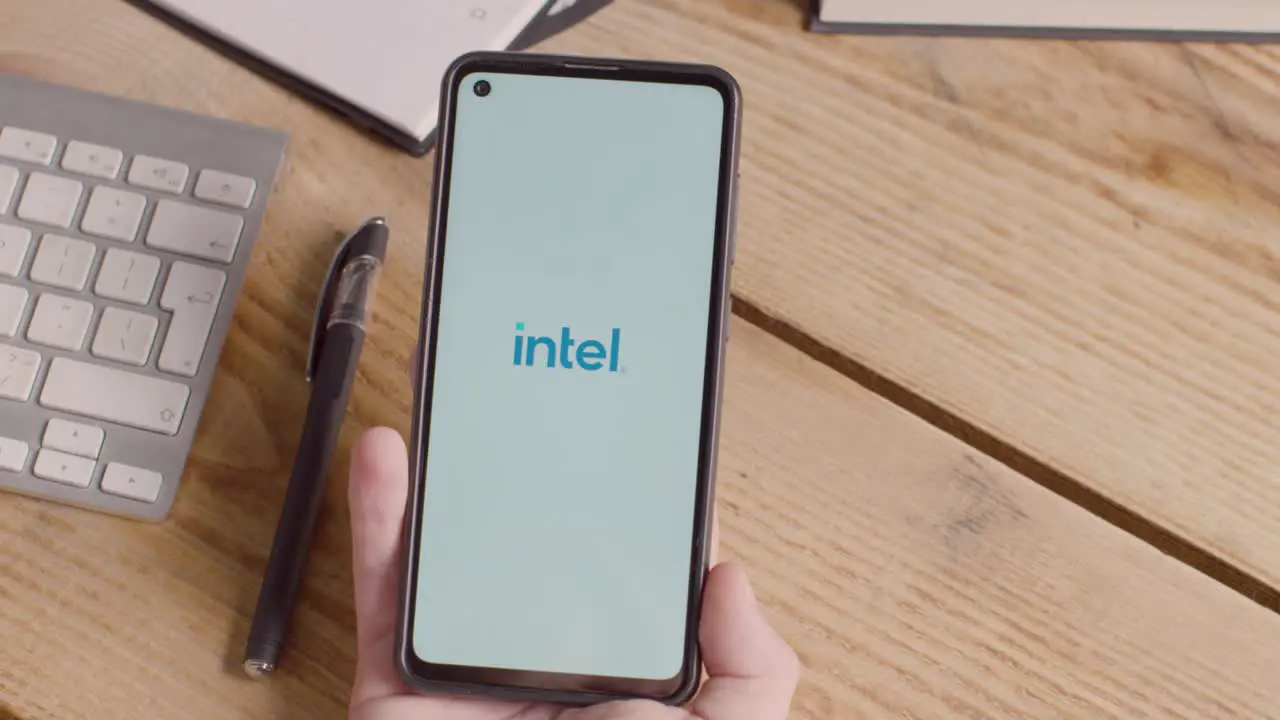 Person Holding Mobile Phone On Desk With Screen Showing Brand Logos For Microsoft Intel IBM nVidia Adobe and Qualcomm