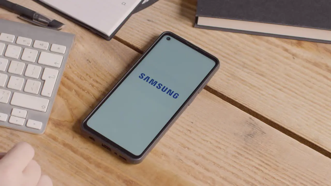 Mobile Phone On Desk With Screen Showing Brand Logos For Apple And Samsung