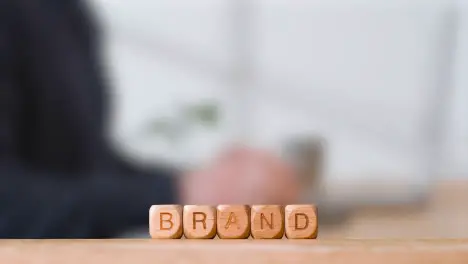 Business Concept Wooden Letter Cubes Or Dice Spelling Brand With Office Person Using Mobile Phone In Background