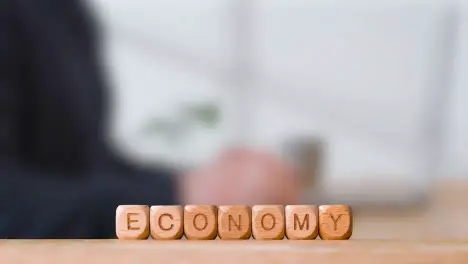 Business Concept Wooden Letter Cubes Or Dice Spelling Economy With Office Person Using Mobile Phone In Background