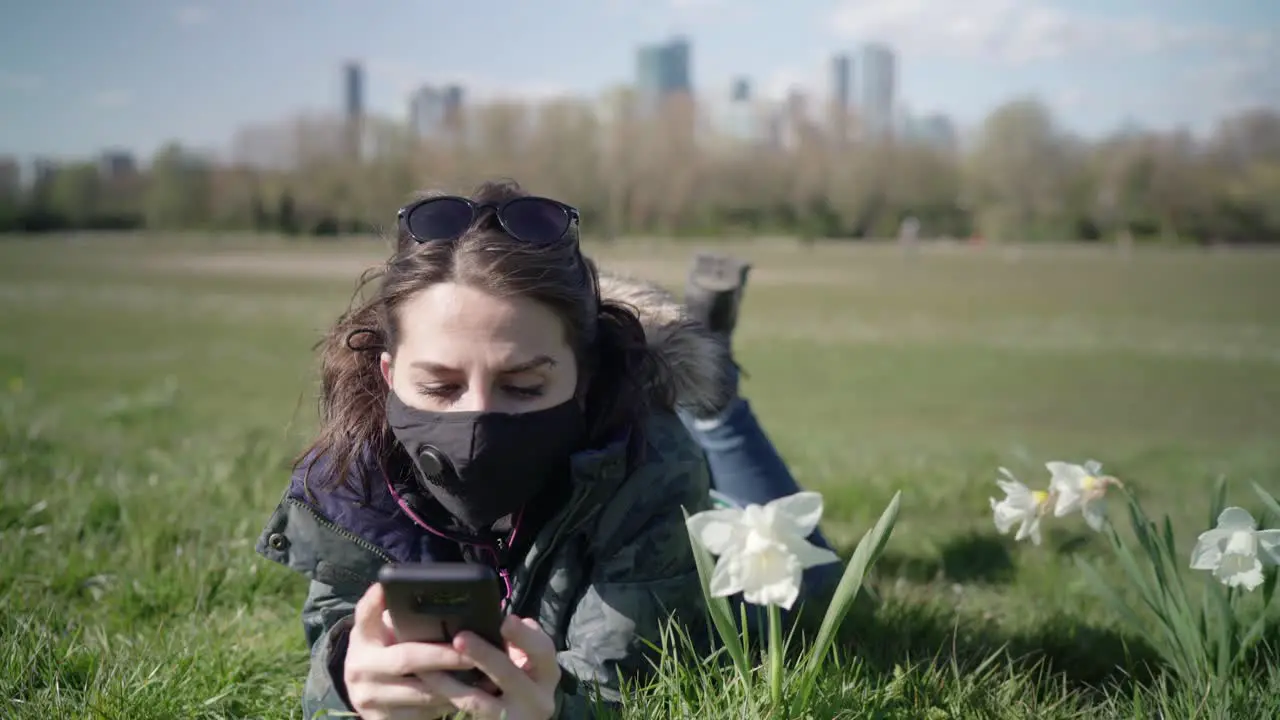 Woman wearing a black face mask lying on the grasses and playing with her phone in the park during a summer day with skyscrapers in the horizon