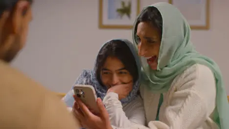 Muslim Mother And Daughter Laughing At Mobile Phone Sitting Around Muslim Family Table At Home