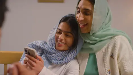 Muslim Mother And Daughter Laughing At Mobile Phone Sitting Around Muslim Family Table At Home 1