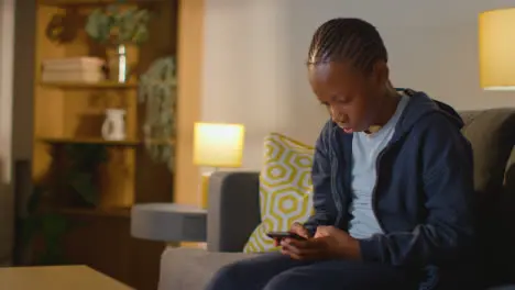 Boy At Home Sitting On Sofa In Evening Looking At Mobile Phone 