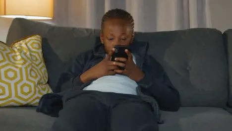 Boy At Home Sitting On Sofa In Evening Using Mobile Phone