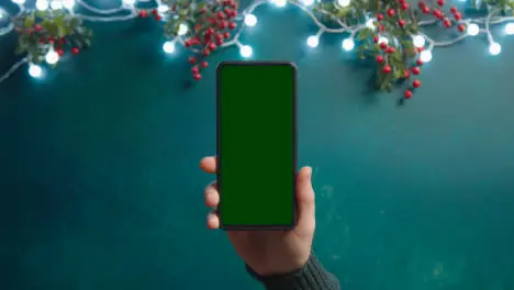 Overhead Shot Of Person Holding Green Screen Mobile Phone With Christmas Decorations And Holly Berries