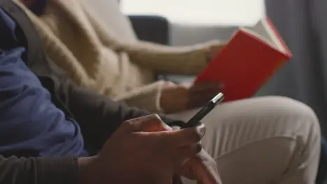 Close Up Of Couple At Home Sitting On Sofa In Lounge With Woman Reading Book And Man Looking At Mobile Phone