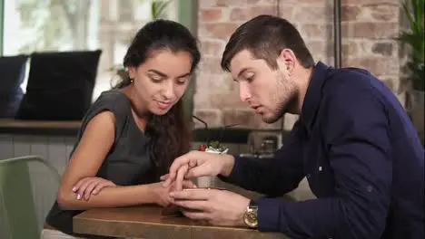 Cute young couple holding hands and using phone sitting at the wooden table in coffee shot Romantic date