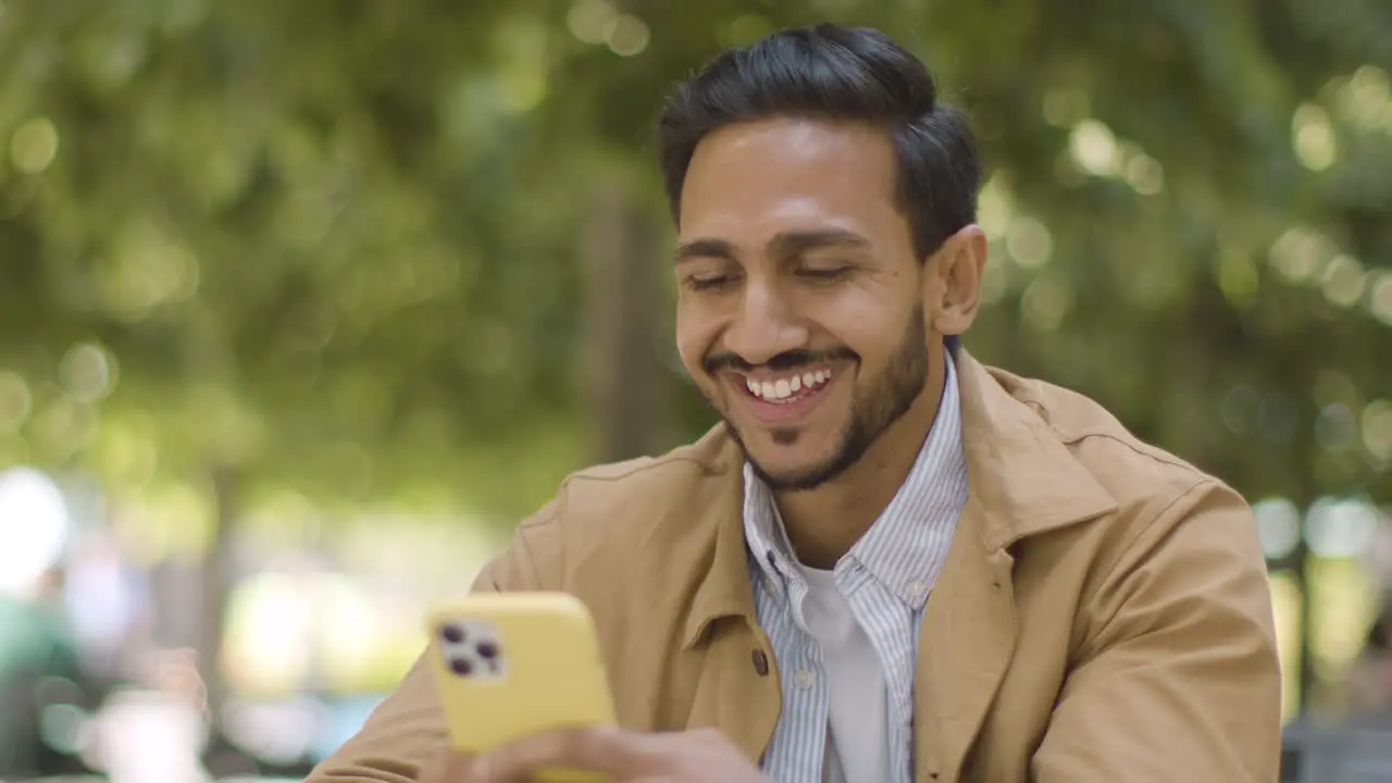 Muslim Man Sitting At Outdoor Table On City Street Sending Text Message To Date On Mobile Phone 1