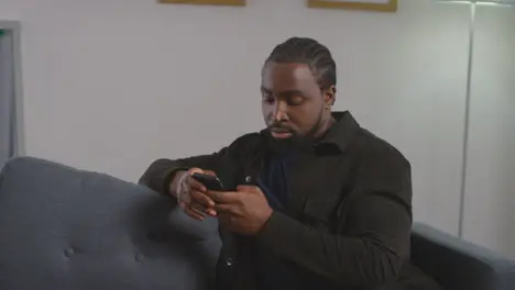 Close Up Of Young Man Relaxing At Home Sitting On Sofa Scrolling Through Mobile Phone