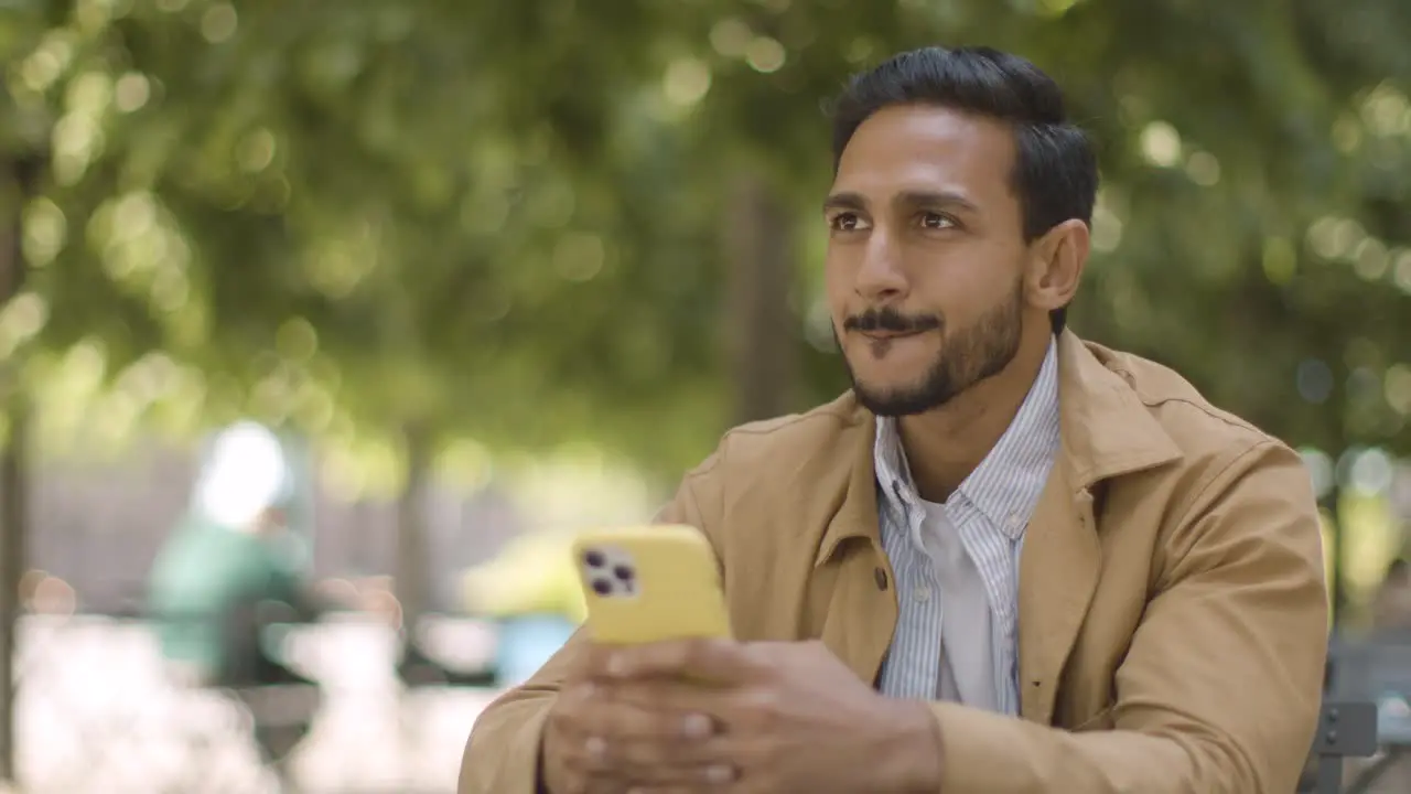 Muslim Man Sitting At Outdoor Table On City Street Sending Text Message To Date On Mobile Phone