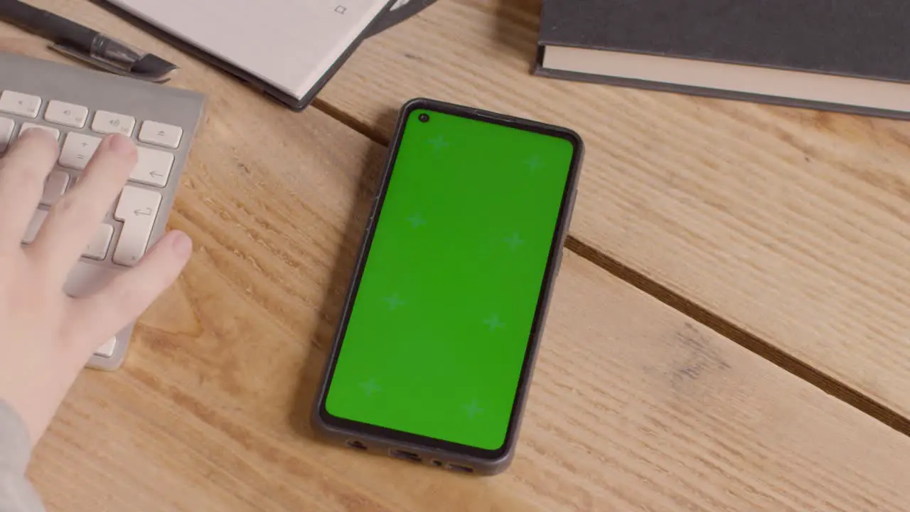 Close Up Of Green Screen Mobile Phone On Desk With Hand Typing On Computer Keyboard 1