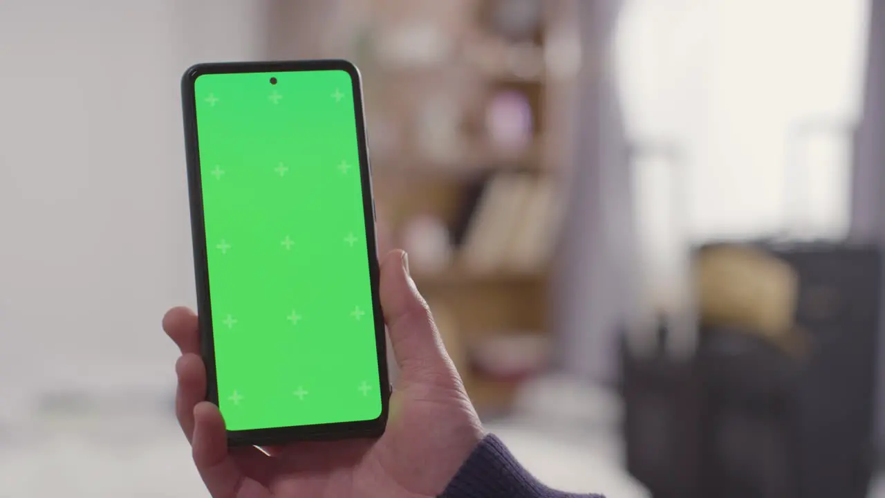 Person At Home Holding Green Screen Mobile Phone With Suitcase Packed For Summer Holiday In Background