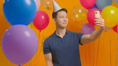 Studio Portrait Of Man Taking Selfie Wearing Party Hat Celebrating Birthday Surrounded By Balloons