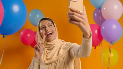 Studio Portrait Of Woman Taking Selfie Wearing Hijab Celebrating Birthday Party Surrounded By Balloons