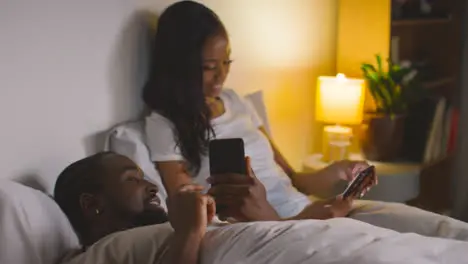 Smiling Couple At Home At Night Both Looking At Mobile Phones In Bed