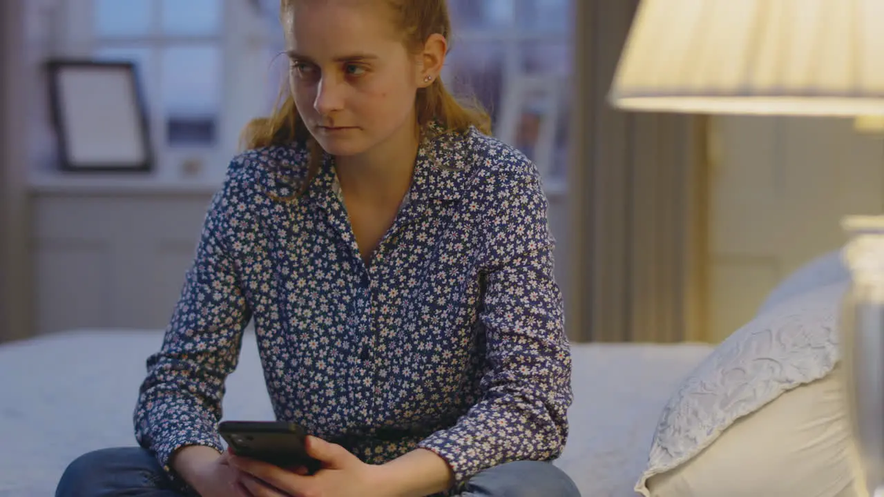 Depressed young woman at home sitting in bedroom in evening with mobile phone looking sad- shot in slow motion