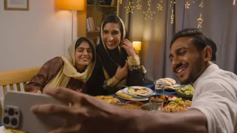 Muslim Family Sitting Around Table At Home With Food For Meal Celebrating Eid Posing For Selfie