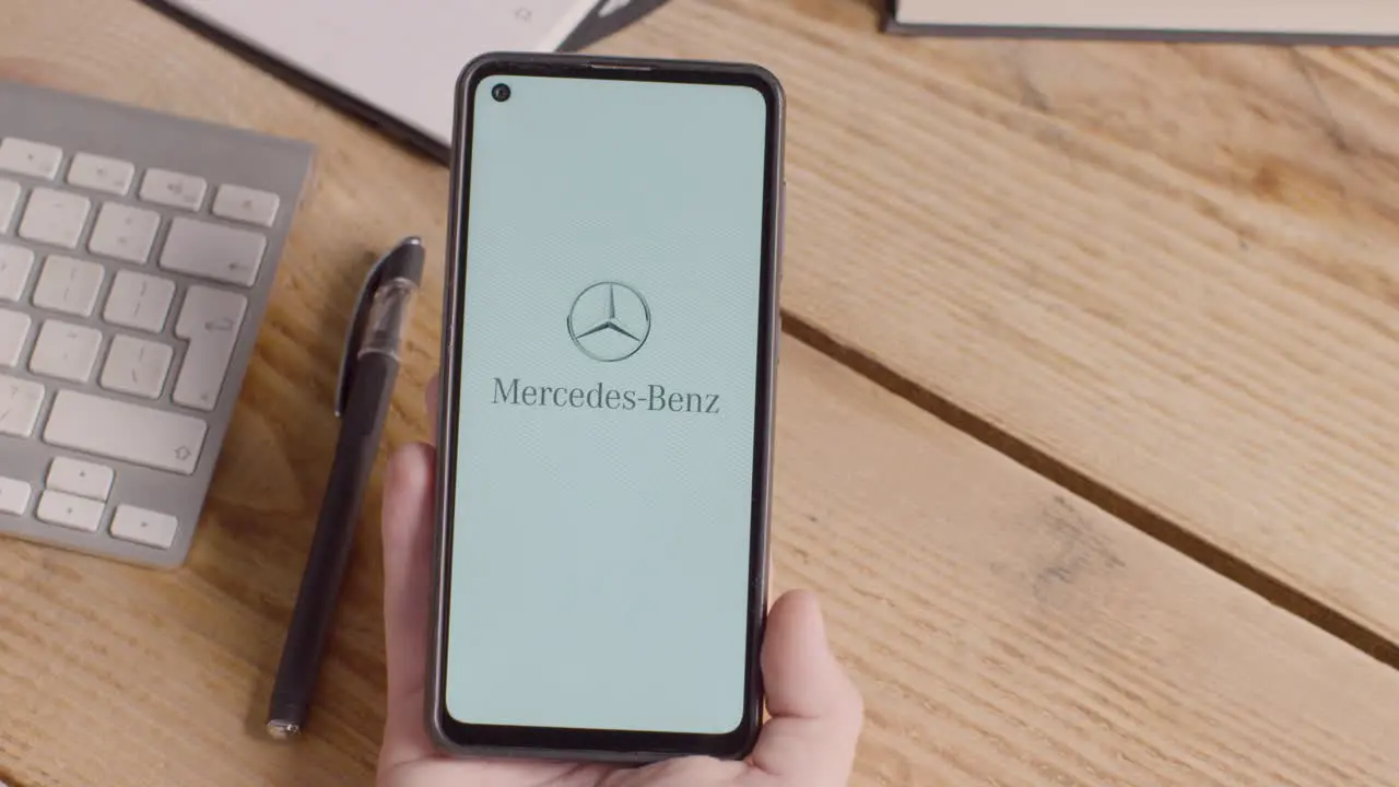 Person Holding Mobile Phone On Desk With Screen Showing Brand Logos For Tesla Mercedes Benz BMW and Toyota