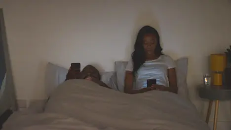 Couple At Home At Night Both Looking At Their Mobile Phones In Bed