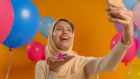 Studio Portrait Of Woman Taking Selfie Wearing Hijab Celebrating Birthday Blowing Paper Confetti Surrounded By Balloons