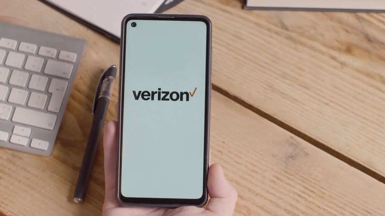 Person Holding Mobile Phone On Desk With Screen Showing Brand Logos For Tencent Verizon AT&T Spectrum Xfinity and Apple