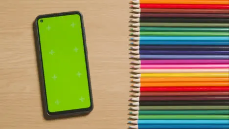 Coloured Pencils In A Line On Wooden Background With Person Scrolling On Green Screen Mobile Phone