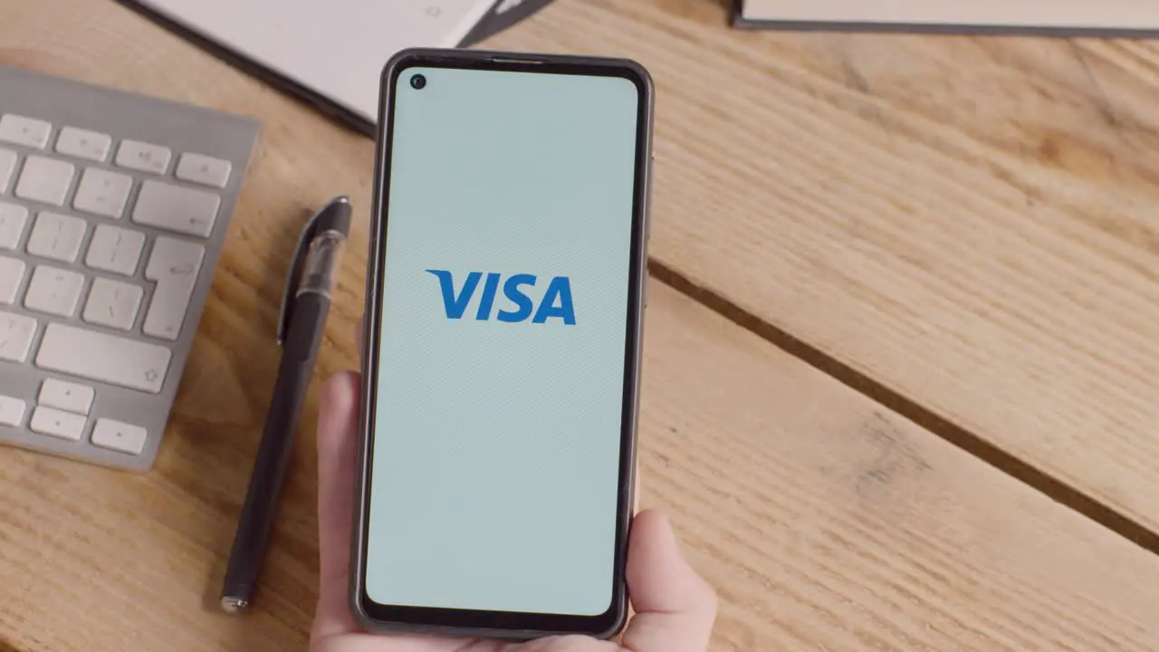 Person Holding Mobile Phone On Desk With Screen Showing Brand Logos For Qualcomm Visa Mastercard American Express and Paypal