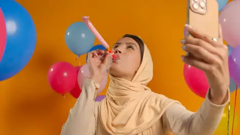 Studio Portrait Of Woman Taking Selfie Wearing Hijab Celebrating Birthday With Party Blower Surrounded By Balloons