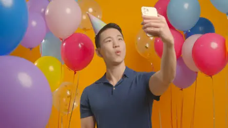 Studio Portrait Of Man Taking Selfie Wearing Party Hat Celebrating Birthday Surrounded By Balloons 1