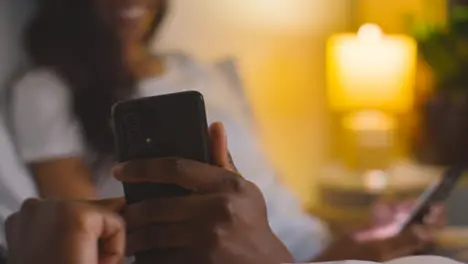 Close Up Of Smiling Couple At Home At Night Both Looking At Mobile Phones In Bed