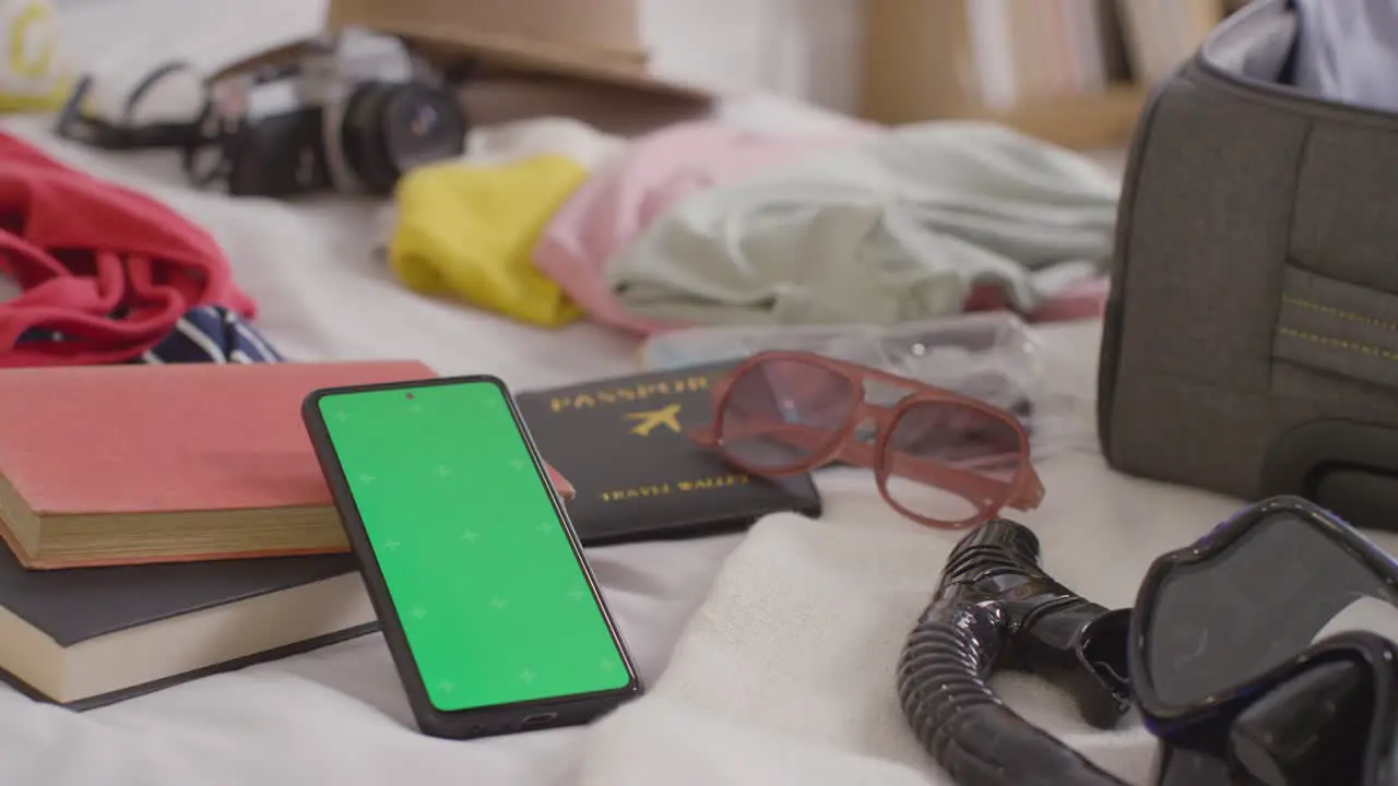 Green Screen Mobile Phone With Open Suitcase On Bed At Home Being Packed For Summer Holiday 