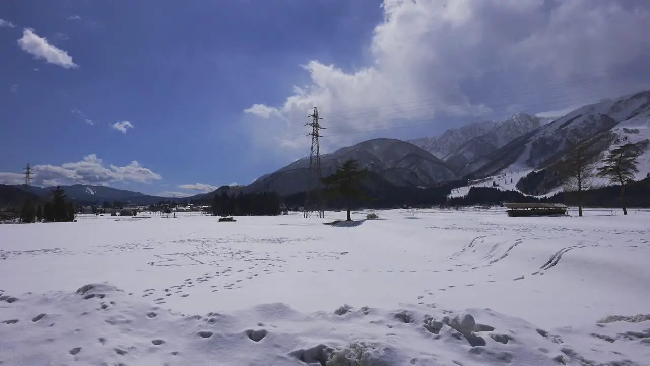 Amidst the captivating winter scenery of Hakuba Japan a shot captures the presence of electrical pylons towering against the cloudy sky while a car gracefully cross the snowy field in the frame