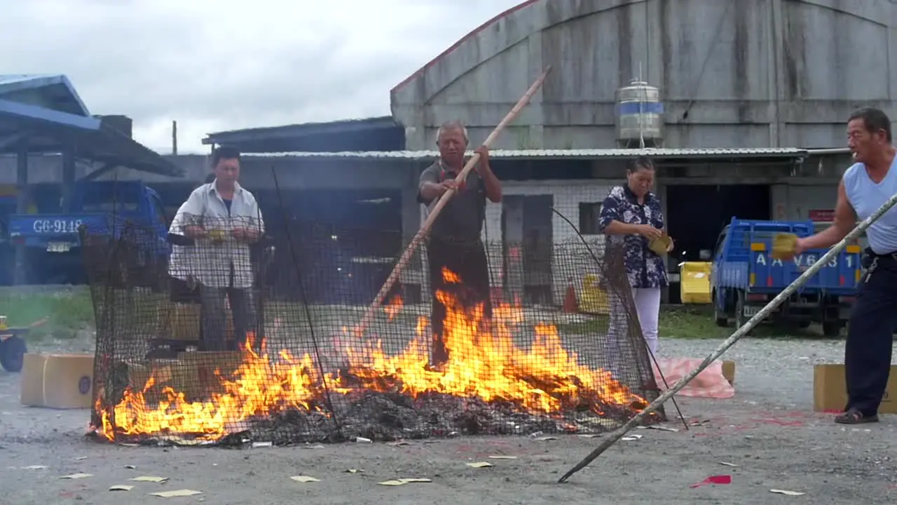 Asian man using large bamboo stick to stir burning paper money on fire during spiritual ceremony to appease ancestors and ghosts