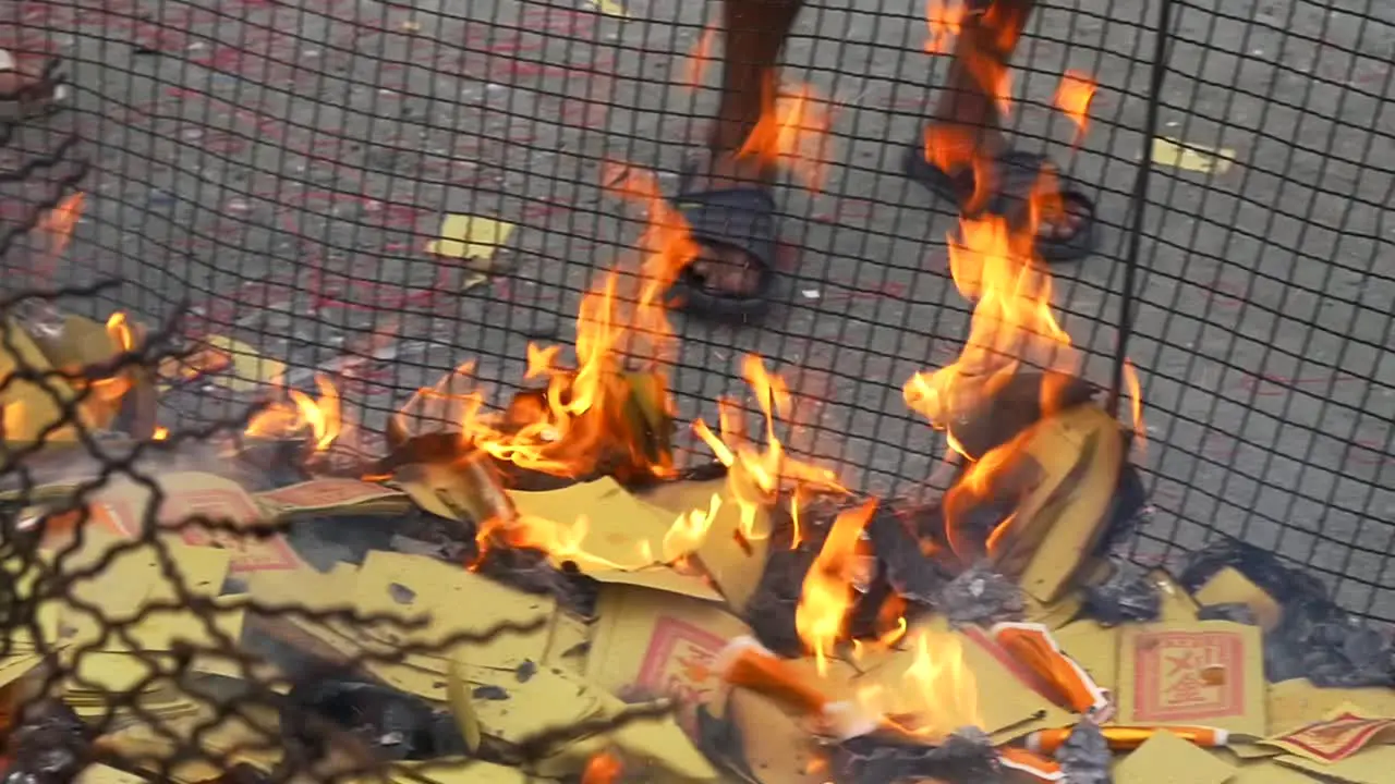 Paper money on fire inside caged fire pit with persons feet in background during spiritual ceremony to appease ancestors and ghosts filmed in slow motion