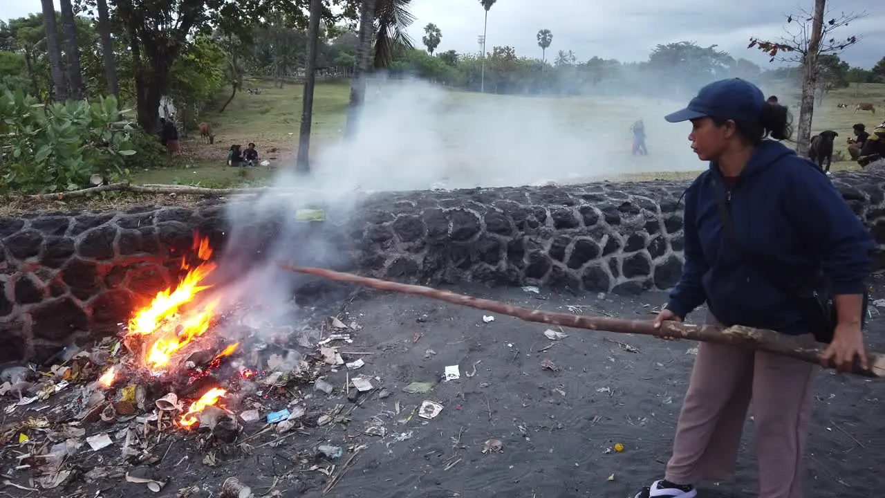 Woman With a Stick Burns Plastic Trash creating Smoke and Fire Asian Beach at Bali Indonesia Toxic Fumes Pollution