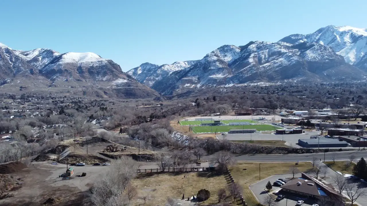 Drone flying high over small town America with high school football field roads and beautiful snow capped mountains on a sunny clear winter day