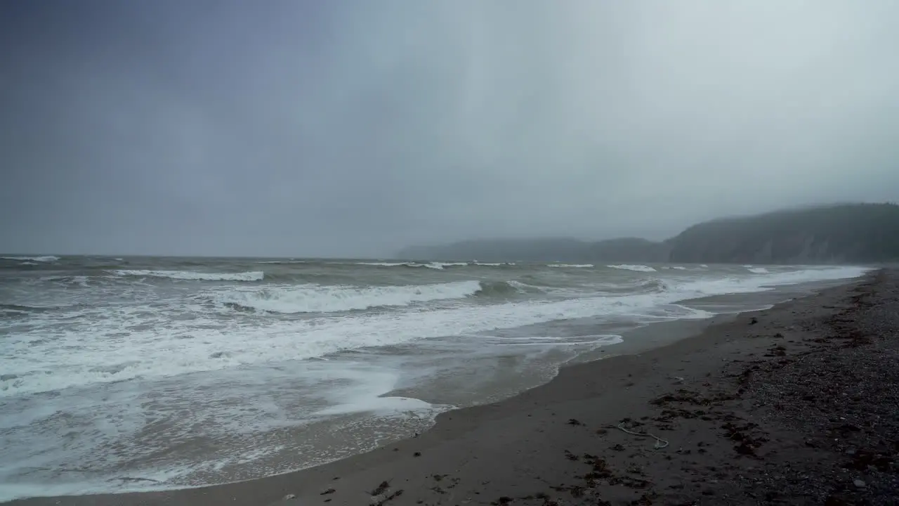 Waves Roll in Creating Sea Foam on an Empty Beach with Fog Covered Sea Cliffs in the Background on a Depressing Cold Wintery Morning Perce Quebec