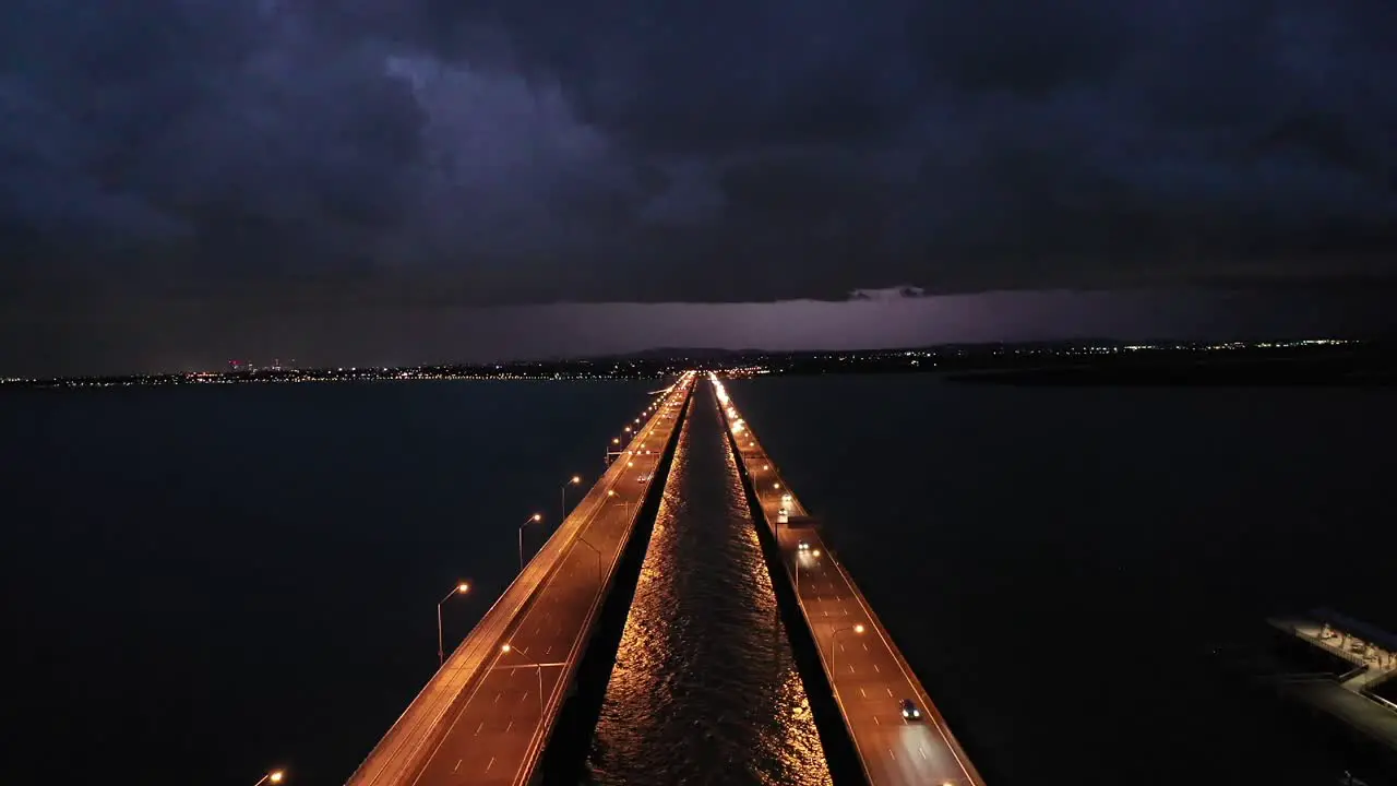 Stationary drone shot of Bridge and Ocean with Storm in the Distance lightning striking the town in distance taken at night