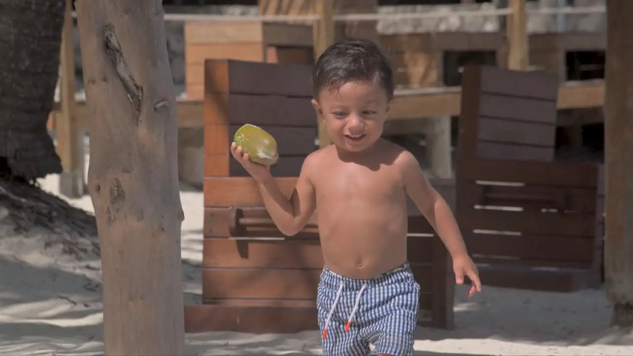 Young latin baby boy walking at the beach on a sunny day wearing his swimsuit shorts and carrying a coconut