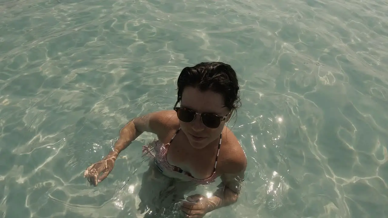 A beautiful girl in her 20s in swimwear and sunglasses waving at the camera and smiling